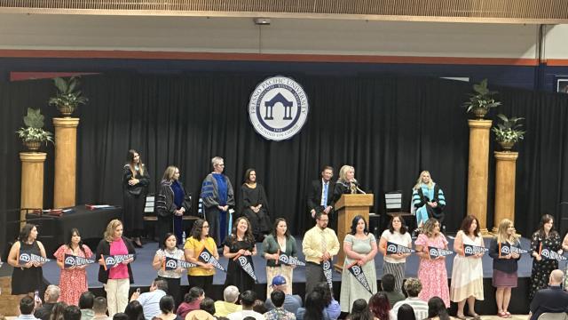 A row of teacher credential recipients stands before the crowd at the FPU Teacher Credential Celebration. Behind them onstage are FPU faculty.