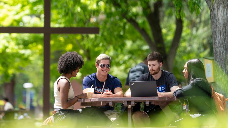 Students sitting outside around a table on campus with a cross in the background