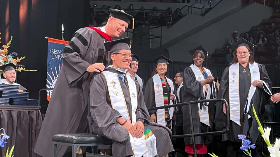 Brian Ross, D.Min., associate professor of pastoral ministries at Fresno Pacific Biblical Seminary, places a master’s hood on a graduate of the M.A. in Ministry, Leadership & Culture program, who is seated on a bench as Brian puts on the hood from the back. Other graduates look on.