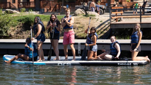 Seven male and female students standing and smiling on a board on the water at Hume Lake.